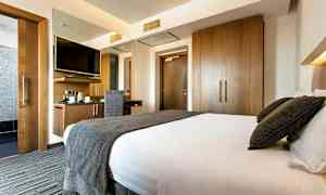 Ett hotellrum med en stor säng, en skrivbordsplats med stol och väggmonterad TV, samt garderober i trä. (AI-genererad text)