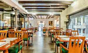 En interiör av en restaurang med tomma bord och stolar, träbjälkar i taket och stora fönster mot en gata. (AI-genererad text)