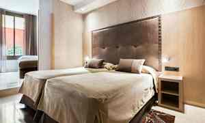 Ett hotellrum med två enkelsängar bredvid varandra och en huvudsäng i mörkt material. Det finns ett litet sängbord och en stor fönsteryta som släpper in naturligt ljus. (AI-genererad text)