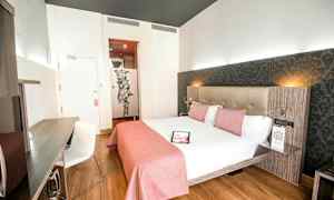 Ett ljust hotellrum med en dubbelsäng bäddad med vita lakan och rosa sängkläder, ett skrivbord med en stol och en dekorativ vägg bakom sängen. (AI-genererad text)