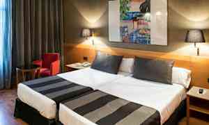 Ett hotellrum med två enkelsängar, varav båda är bäddade med svartvita överkast. På väggen hänger en färgglad tavla, och rummet har även ett litet bord, en röd fåtölj och två vägglampor. (AI-genererad text)