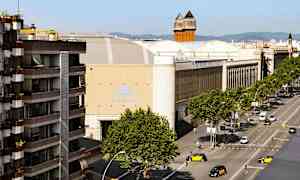 En gata med träd och fordon framför stora byggnader, inklusive en med skylten 'Fira de Barcelona'. (AI-genererad text)