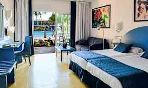 Ett hotellrum med en stor säng, sittgrupp och utsikt över en pool genom glasdörrar. (AI-genererad text)