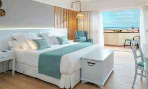 Ett ljust hotellrum med en stor dubbelsäng, stol och ett bord, samt en balkong med utsikt över havet. (AI-genererad text)