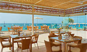 Bild av en restaurang med flätade stolar och glasbord, belägen vid havet med utsikt över en blå vattenhorisont. (AI-genererad text)