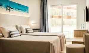 Ett hotellrum med två enkelsängar, vita väggar och ett fönster som vetter mot en balkong. En målning pryder väggen ovanför sängarna. (AI-genererad text)