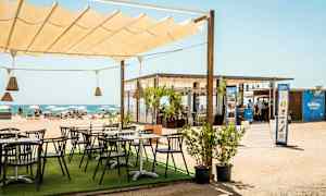 En strandbar med uteservering under ett segeltak, med utsikt över stranden och havet. (AI-genererad text)