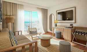 Ett ljust och modernt hotellrum med utsikt över havet. Det finns en soffa, ett soffbord, en stol och en TV på väggen. (AI-genererad text)