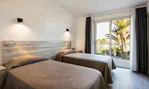 Två enkelsängar i ett ljust rum med stora fönster mot en balkong och utsikt över palmer. (AI-genererad text)