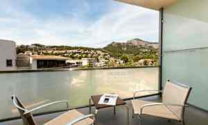 Balkong med två stolar, ett bord och en glasskärm med utsikt över en bergig och urban miljö. (AI-genererad text)