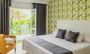 Ett hotellrum med en dubbelsäng och färgstarkt mönstrat tapet. Balkongdörrarna står öppna och visar en grönskande utsikt. (AI-genererad text)