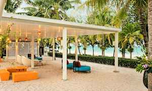 En täckt uteplats med sittmöbler på en sandstrand, omgiven av palmer och med utsikt över havet. (AI-genererad text)