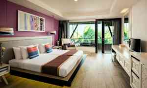 Ett hotellrum med en bred bäddad säng, dekorativa kuddar, ett skrivbord med en TV och stora glasdörrar som vetter mot en balkong med utsikt över palmer. (AI-genererad text)