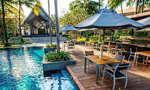 En utomhuspool omgiven av ett trädäck med bord och stolar. Palmer och tropiska växter omgärdar området. (AI-genererad text)