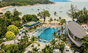 En vy av en semesterort med en stor pool, omgiven av grönska och palmer. I bakgrunden finns en strand och en vik med segelbåtar förankrade. (AI-genererad text)