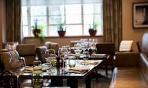 Ett restaurangområde med dukade bord, glas och servetter. I bakgrunden finns fönster med växter. (AI-genererad text)