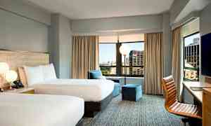 Ett hotellrum med två dubbelsängar, stora fönster med utsikt över staden, en fåtölj och ett skrivbord med en stol. (AI-genererad text)