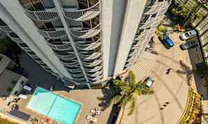 Ett höghus sett uppifrån med balkonger som vetter utåt. Nedanför finns en pool, solstolar och parkerade bilar. (AI-genererad text)