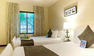 Ett hotellrum med två breda sängar, varsin kudde och pläd. Ett fönster med draperier och utsikt över träd. (AI-genererad text)