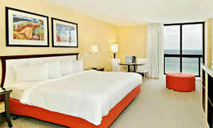 Ett hotellrum med en stor säng som har vit sängklädsel och en orange sängbotten. Rummet har två tavlor ovanför sängen, ett runt bord med två stolar, en orange sittpuff och ett fönster med havsutsikt. (AI-genererad text)