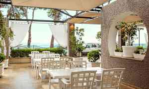 Utomhusområde med vita bord och stolar under ett tak, omgivna av grön växtlighet och havsutsikt. (AI-genererad text)