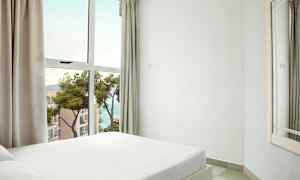 Ett sovrum med en vit säng nära ett stort fönster med utsikt över havet och närliggande byggnader. (AI-genererad text)