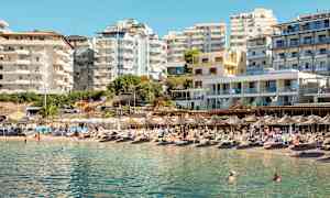 En strand med solstolar och parasoller framför flera modern byggnader. (AI-genererad text)