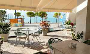 Utomhusservering med bord och stolar framför glasvägg, hav och träd i bakgrunden. (AI-genererad text)