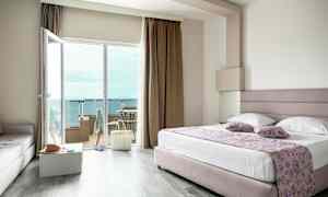 Ett hotellrum med en stor säng, glasdörrar till en balkong och utsikt över havet. (AI-genererad text)