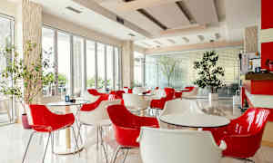 Ett modernt café med röda och vita stolar samt bord, beläget i ett ljust rum med stora fönster och inomhusväxter. (AI-genererad text)