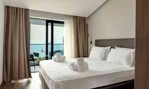 Ett hotellrum med en bäddad säng och utgång till en balkong med havsutsikt. (AI-genererad text)