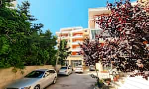 Bilden visar ett bostadshus med parkerade bilar framför och buskar samt träd runtom. (AI-genererad text)