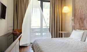 Ett hotellrum med en säng, TV, bord och stol, samt ett fönster med vita gardiner. (AI-genererad text)