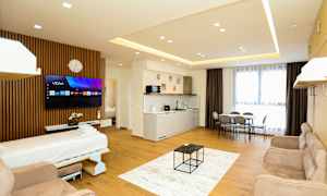 Ett modernt hotellrum med öppen planlösning inkluderande en bäddsoffa, TV, köksområde, matbord och stora fönster med gardiner. (AI-genererad text)