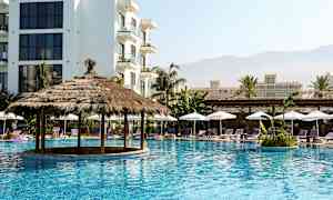 En pool med en halmtakspaviljong omgiven av solstolar och parasoller, med flerbyggnadskomplex i bakgrunden. (AI-genererad text)