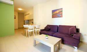 Ett ljust möblerat vardagsrum med en lila soffa, vit soffbord och matplats intill ett kök. (AI-genererad text)