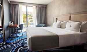 Ett hotellrum med en bäddad dubbelsäng, ett skrivbord med stol och fönster mot balkong. (AI-genererad text)