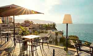 Terrass med matbord och stolar, havsutsikt och byggnader i bakgrunden. (AI-genererad text)