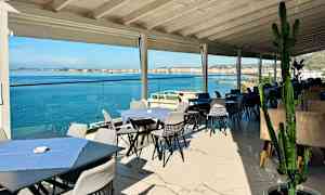 Uteservering med bord och stolar nära havet, med utsikt över vattnet och en stad i bakgrunden. (AI-genererad text)