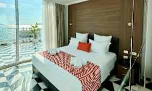 Ett hotellrum med en dubbelbädd täckt med röda och vita textilier, stora glasfönster med havsutsikt. (AI-genererad text)