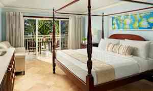 Rummet har en säng med fyra stolpar och är inrett med ljusa möbler. En balkong med utsikt över grönskande palmer syns genom de öppna glasdörrarna. (AI-genererad text)