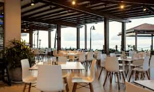 En restaurangterrass med vita stolar och bord under ett trätak. Utsikt över havet bakom. (AI-genererad text)
