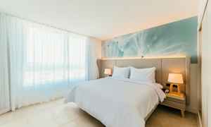 Ljust och modernt inrett sovrum med en dubbelsäng, två sidobord och en stor fönsterpanel med skira vita draperier. (AI-genererad text)