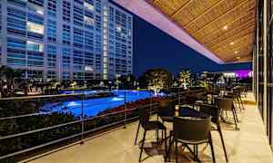 Utsikt över en upplyst pool från en terrass med bord och stolar, omgiven av grönska och med en byggnad i bakgrunden på kvällen. (AI-genererad text)