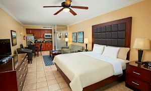 Ett hotellrum med en stor säng, nattduksbord, draperad fåtölj, matplats och ett kök i bakgrunden. (AI-genererad text)