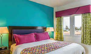 Ett sovrum med en dubbelsäng, färgglada sängkläder och kuddar. Väggarna är turkosa, och rummet har fönster med utsikt över palmer och havet. (AI-genererad text)