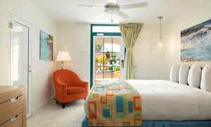 Ett sovrum med ljus inredning. En säng med färgglad överkast, en orange fåtölj, bordslampa och ett fönster med utsikt utåt. (AI-genererad text)