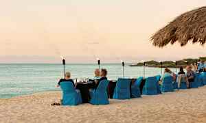 Strand med dukat bord och sittplatser med blå stolsöverdrag, där människor äter och umgås. Havet och himlen syns i bakgrunden. (AI-genererad text)