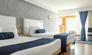 Ett hotellrum med två sängar med vita lakan och blå överkast. Fönstret är täckt med blå gardiner och ljus strömmar in i rummet. (AI-genererad text)