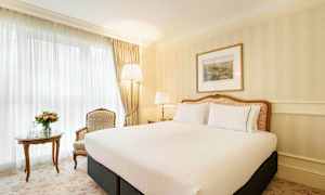 Ett välutrustat sovrum med en stor säng och elegant inredning. Intill sängen står ett bord med en blomsterbukett och en vattenkaraff, bredvid en bekväm stol. (AI-genererad text)